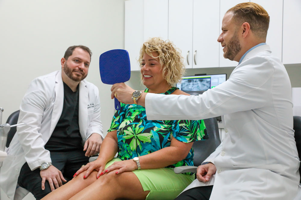 Drs. Alex Planes & Lazaro Gavilla Showing An All-On-4 Dental Implant Patient Their New Smile With A Hand Mirror After Their Procedure